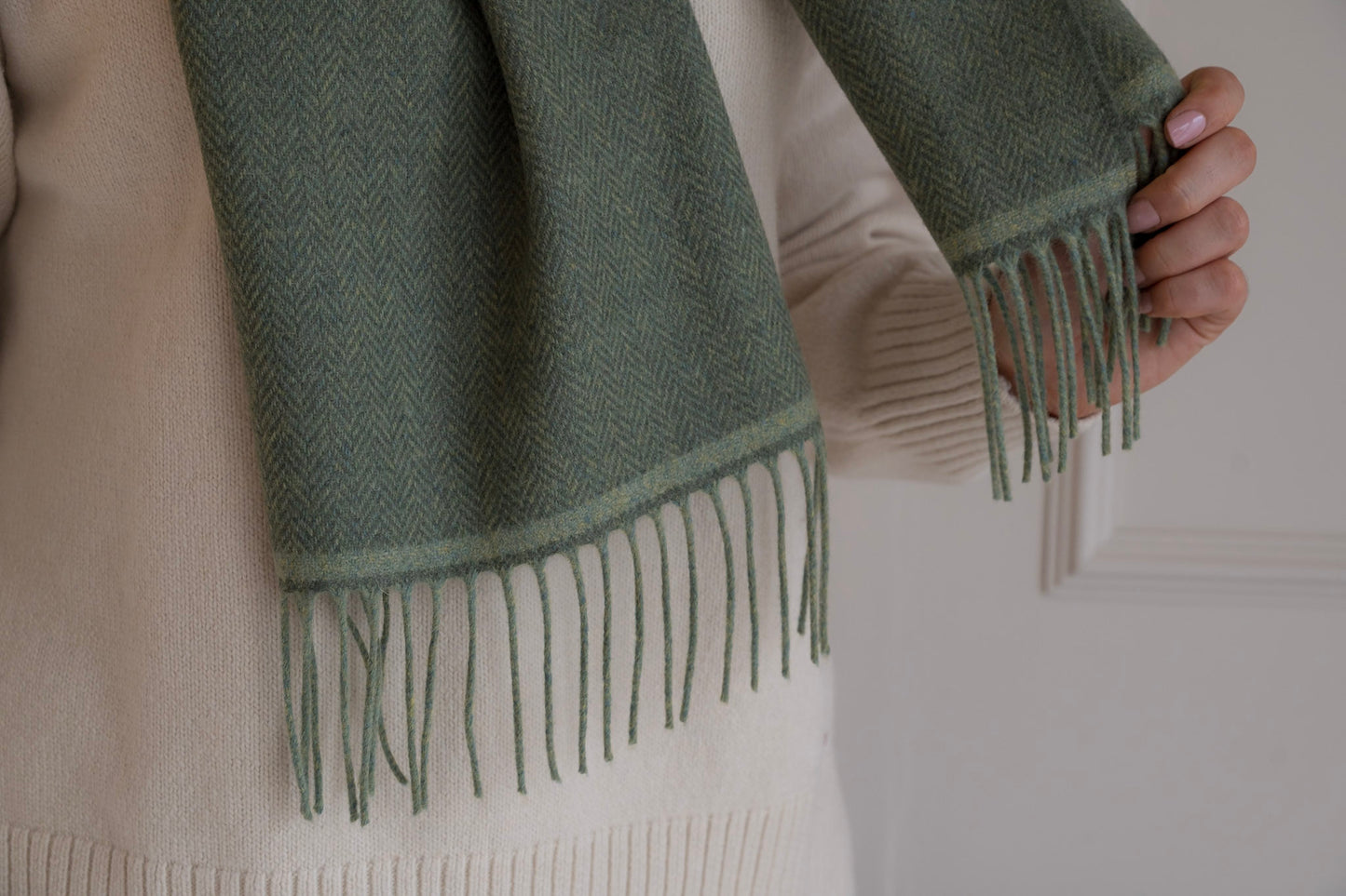 Green scarf with fringes held by a person wearing a beige knitted jumper.