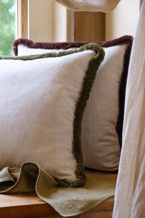 A close-up of a beige-colored herringbone linen cushion with deep olive cotton fringe on a windowsill.