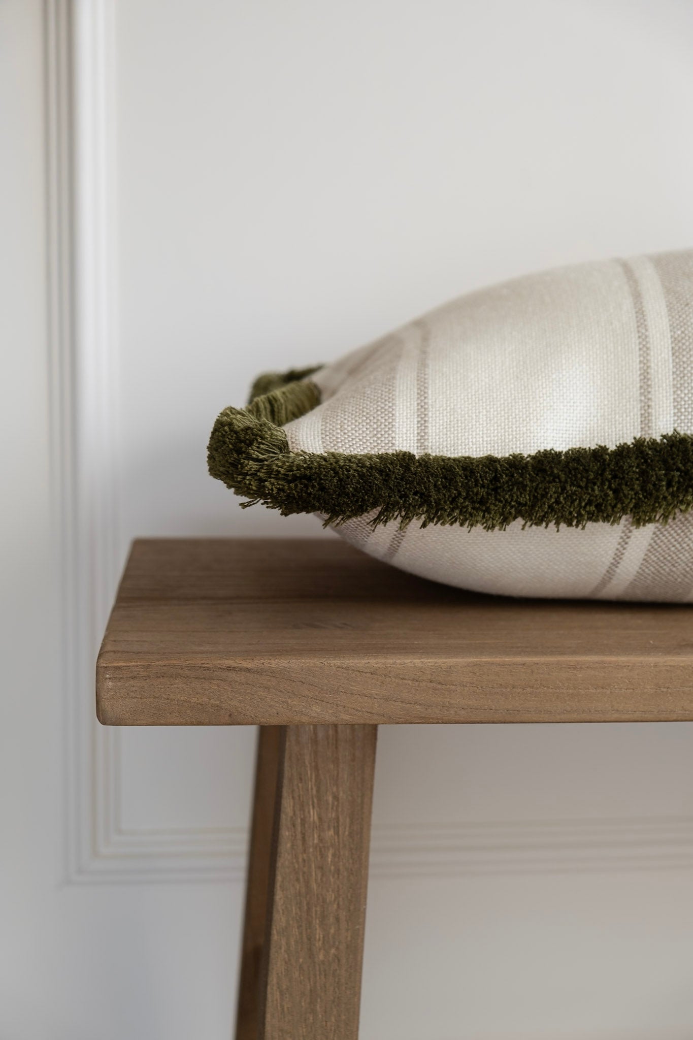A Neutral striped Cushion with a thick Olive Fringe detail. This photo shows the side view of this cushion on a bench
